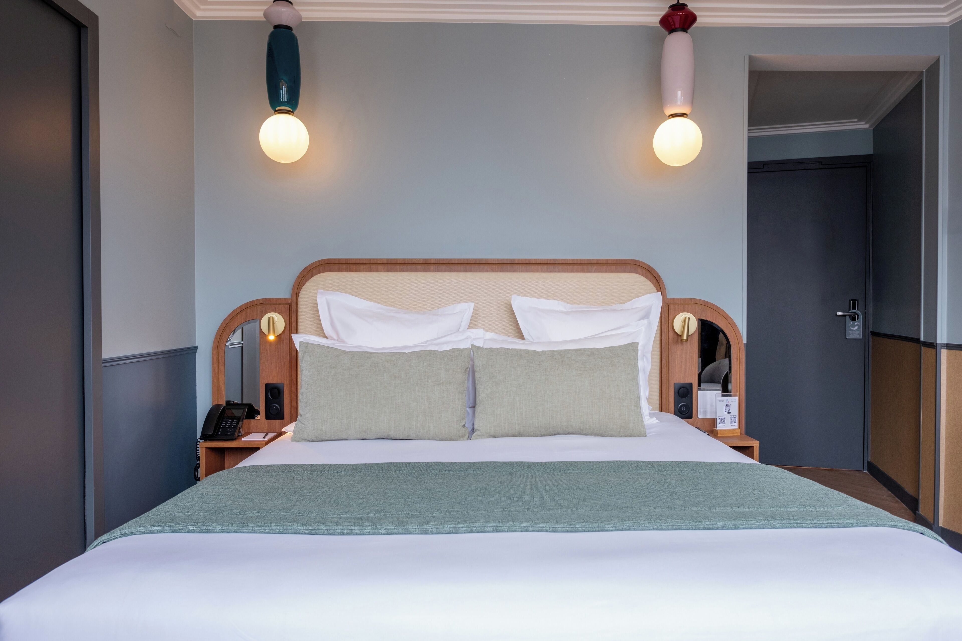 Un lit bien fait, avec du linge de lit blanc et deux oreillers, une table de chevet avec un téléphone et une petite lampe, et deux appliques murales.