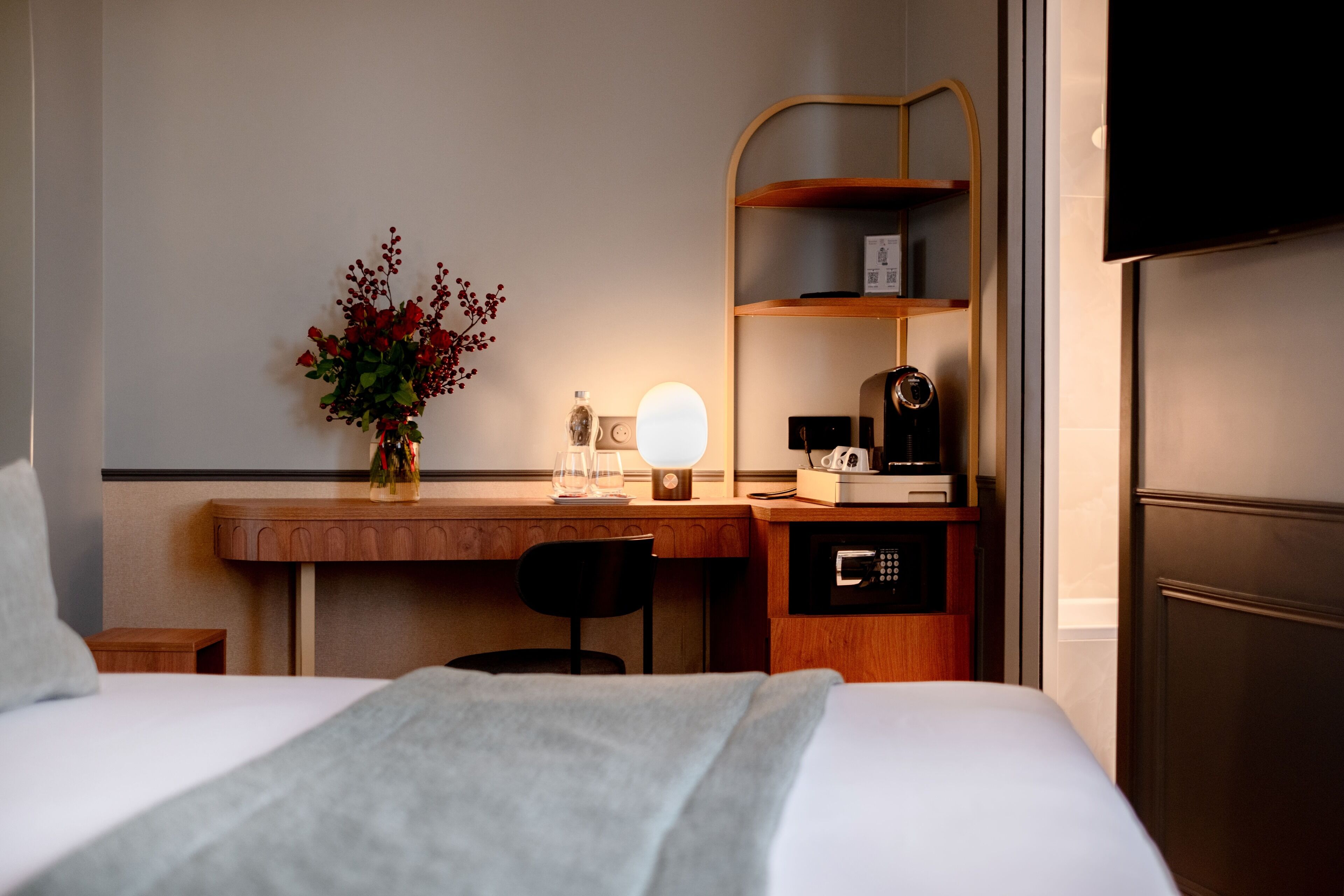 Et hotelværelse med seng, skrivebord med lampe, kaffemaskine og fjernsyn.