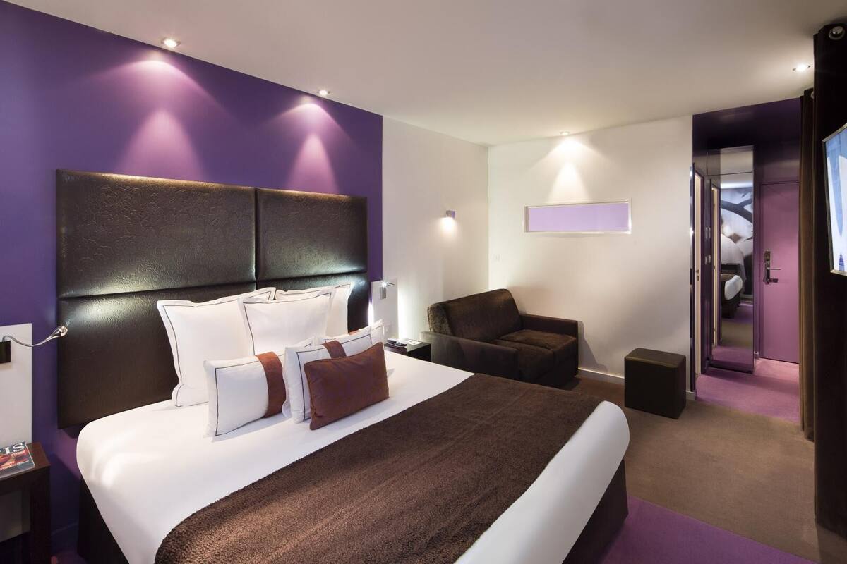 A modern hotel room with a large bed, a dark headboard, a brown armchair, and a small table.
