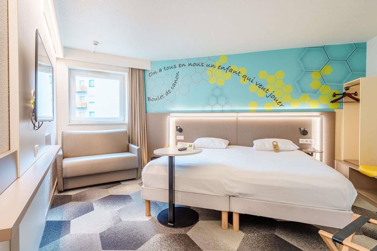 A hotel room with a bed, a chair, a desk, a wall with a hexagonal pattern, and a window.