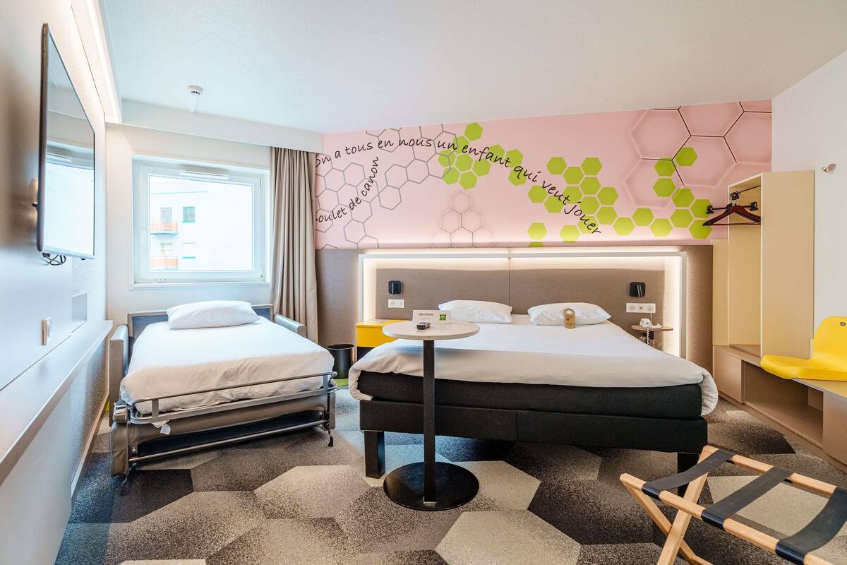 A hotel room with two beds, a small table, a chair, a TV, and a wall with hexagonal patterns.