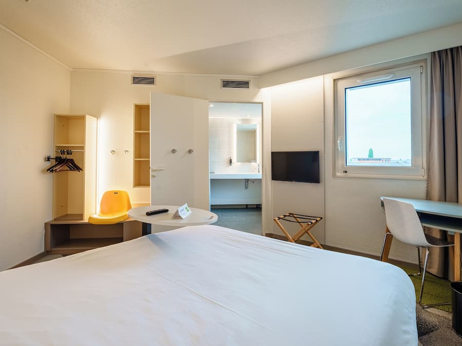 Une chambre d’hôtel avec un grand lit, un bureau avec une chaise, une petite table avec une chaise, un téléviseur fixé au mur et une fenêtre avec des rideaux.