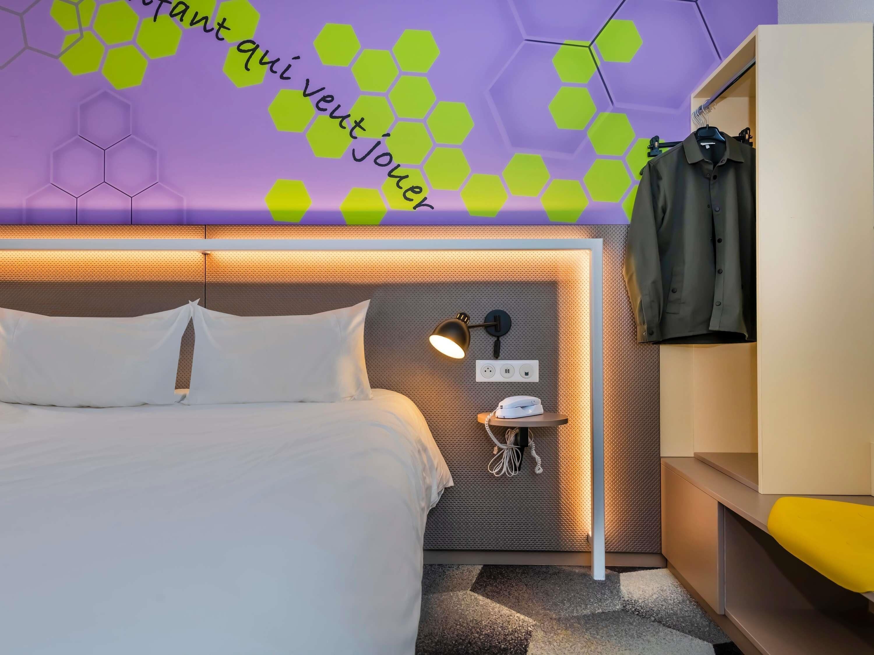 A modern hotel room with a bed, a bedside table with a lamp, a phone, and a coat rack.