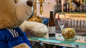 A plush bear wearing a blue shirt sitting at a table with a bottle of wine, a glass, and a bowl of popcorn. - ibis Styles Strasbourg Stade de la Meinau (Strasbourg)