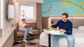 A hotel room with a sofa, a bed, a table with a plate of food, a wall with a hexagonal pattern, and a window. - ibis Styles Strasbourg Stade de la Meinau (Strasbourg)