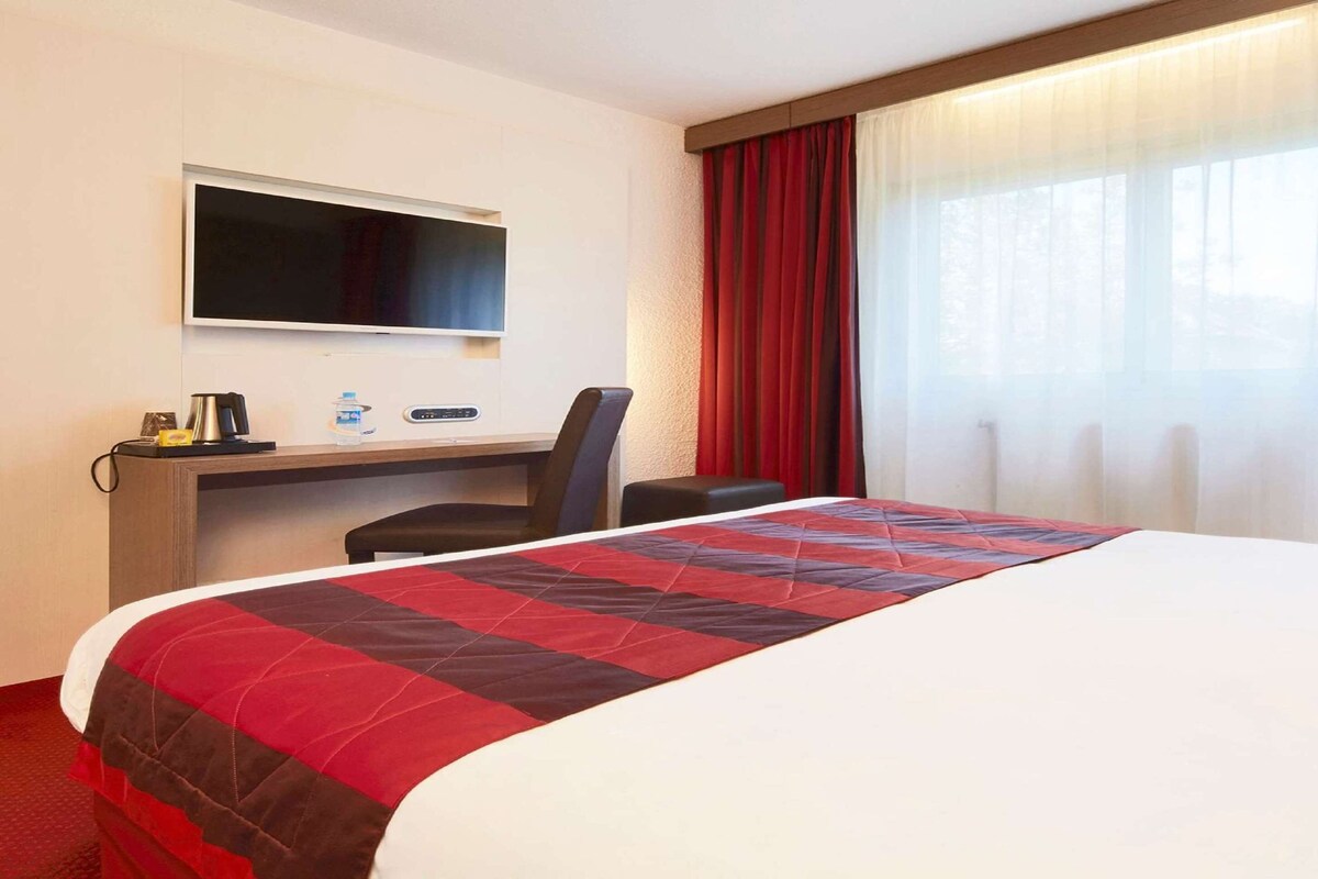 A hotel room with a large bed, a desk with a TV, and a window with red curtains.