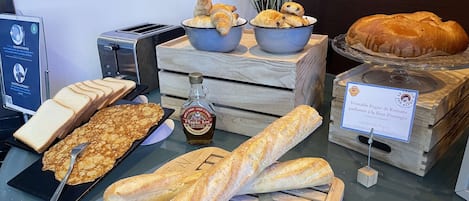 Una exhibición de panes y pasteles diversos, incluyendo croissants, baguettes y una barra de Pigne. También se observa una botella de jarabe Maple Joe y un letrero que indica que el Pigne es hecho a mano y con sabor a azahar.