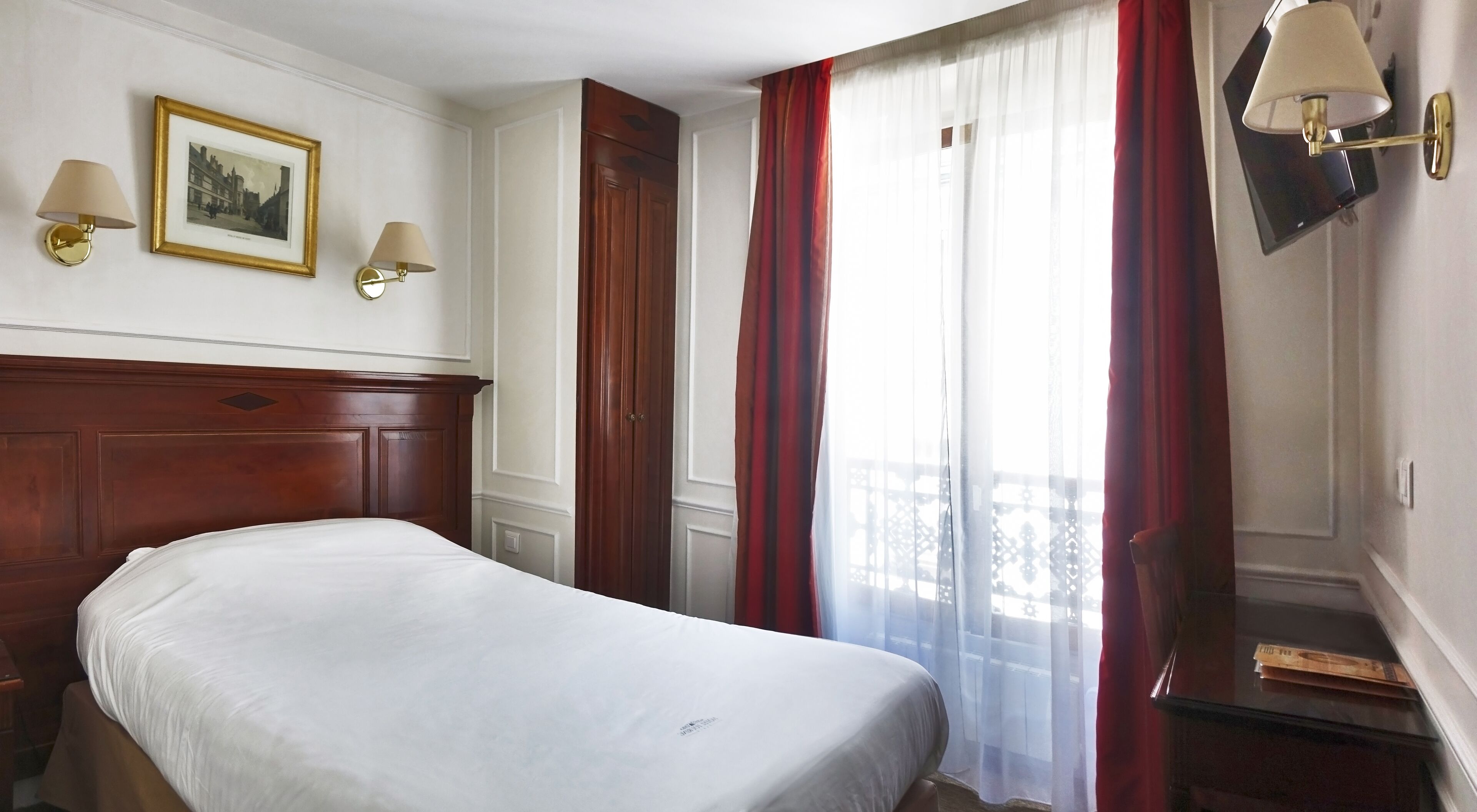 A hotel room with a wooden headboard, a bed with white bedding, two wall-mounted lamps, a framed picture, and a window with red curtains.