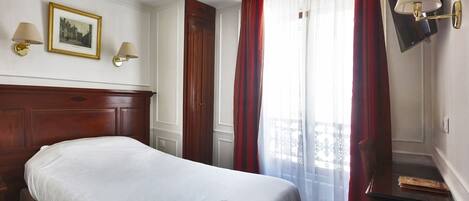 A hotel room with a wooden headboard, a bed with white bedding, two wall-mounted lamps, a framed picture, and a window with red curtains.