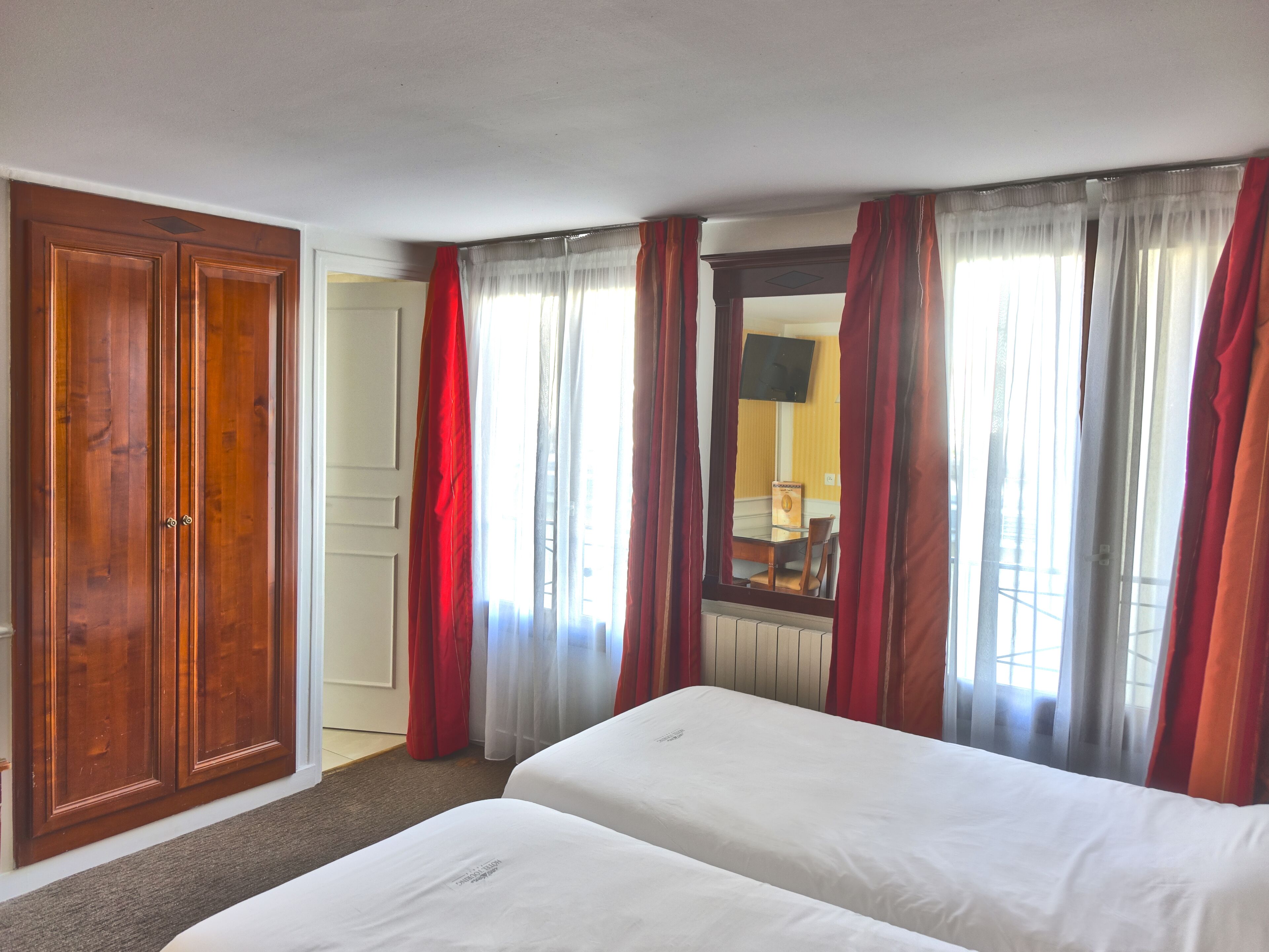 Een hotelkamer met twee bedden, een houten garderobe, een spiegel en rode gordijnen.