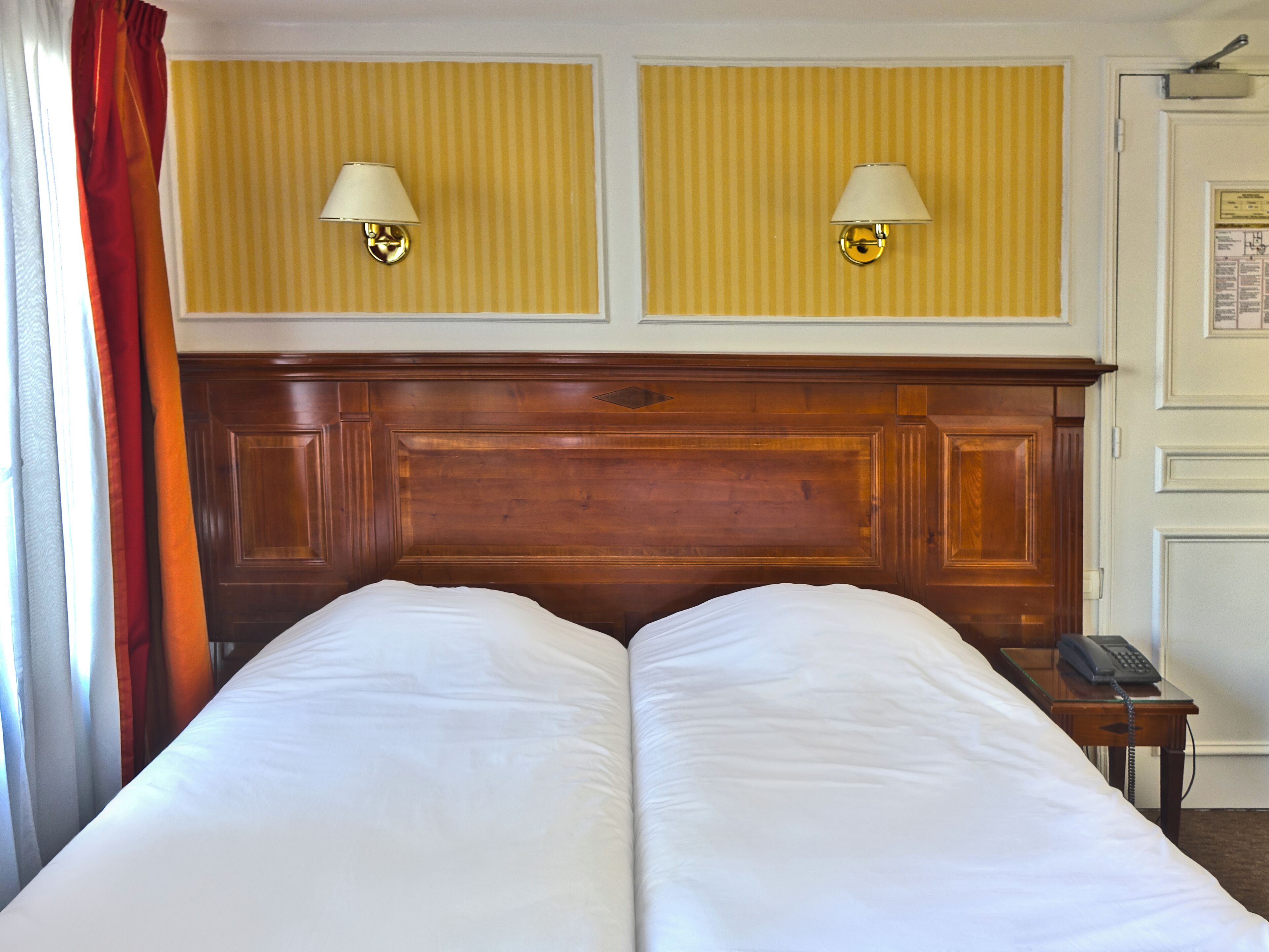 Uma cama de casal com cabeceira de madeira, duas luminárias fixadas na parede e um telefone num criado-mudo.