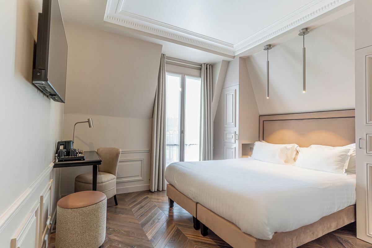 A hotel room with a large bed, a desk with a lamp, a chair, and a waste basket.