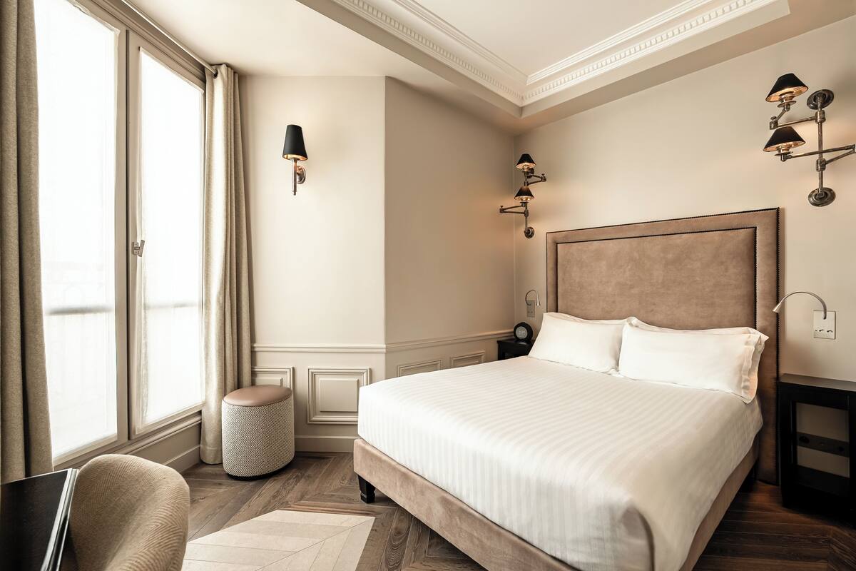 A hotel room with a large bed, a chair, a small round ottoman, and a nightstand with a lamp.