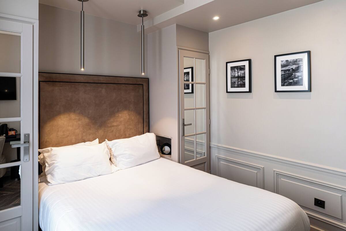 A neatly made bed with white linens, a modern headboard, and a bedside table with a lamp. There are framed pictures on the wall and a mirror reflecting the room.