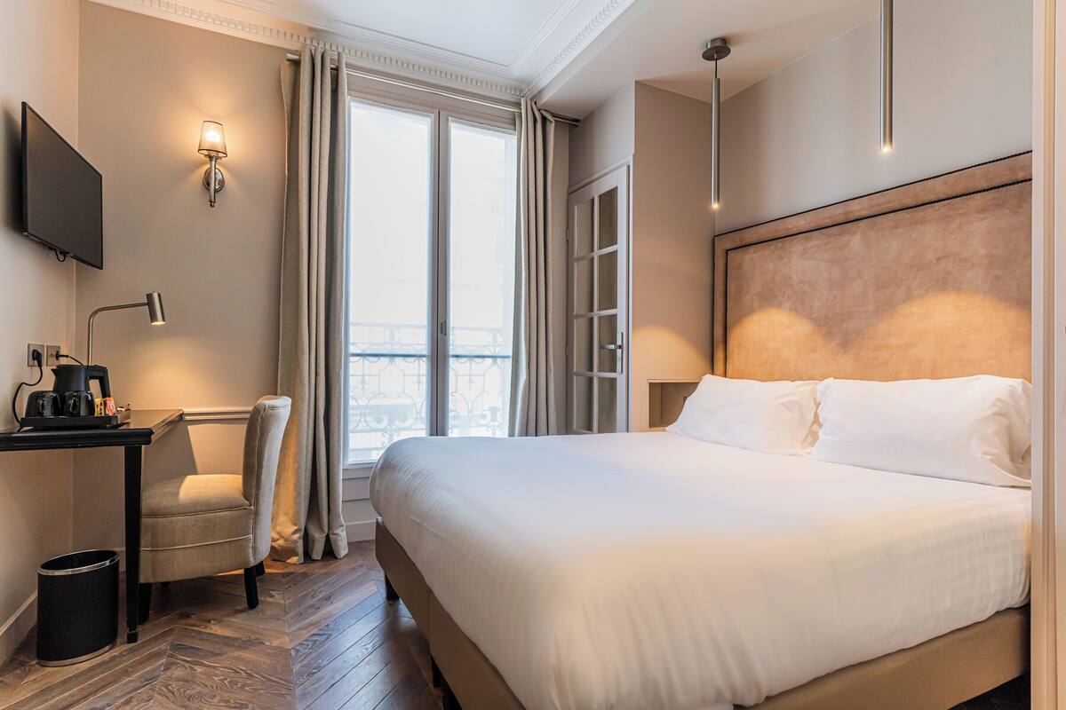 A hotel room with a large bed, a desk with a coffee maker, a chair, a TV mounted on the wall, and a view of the city through the window.