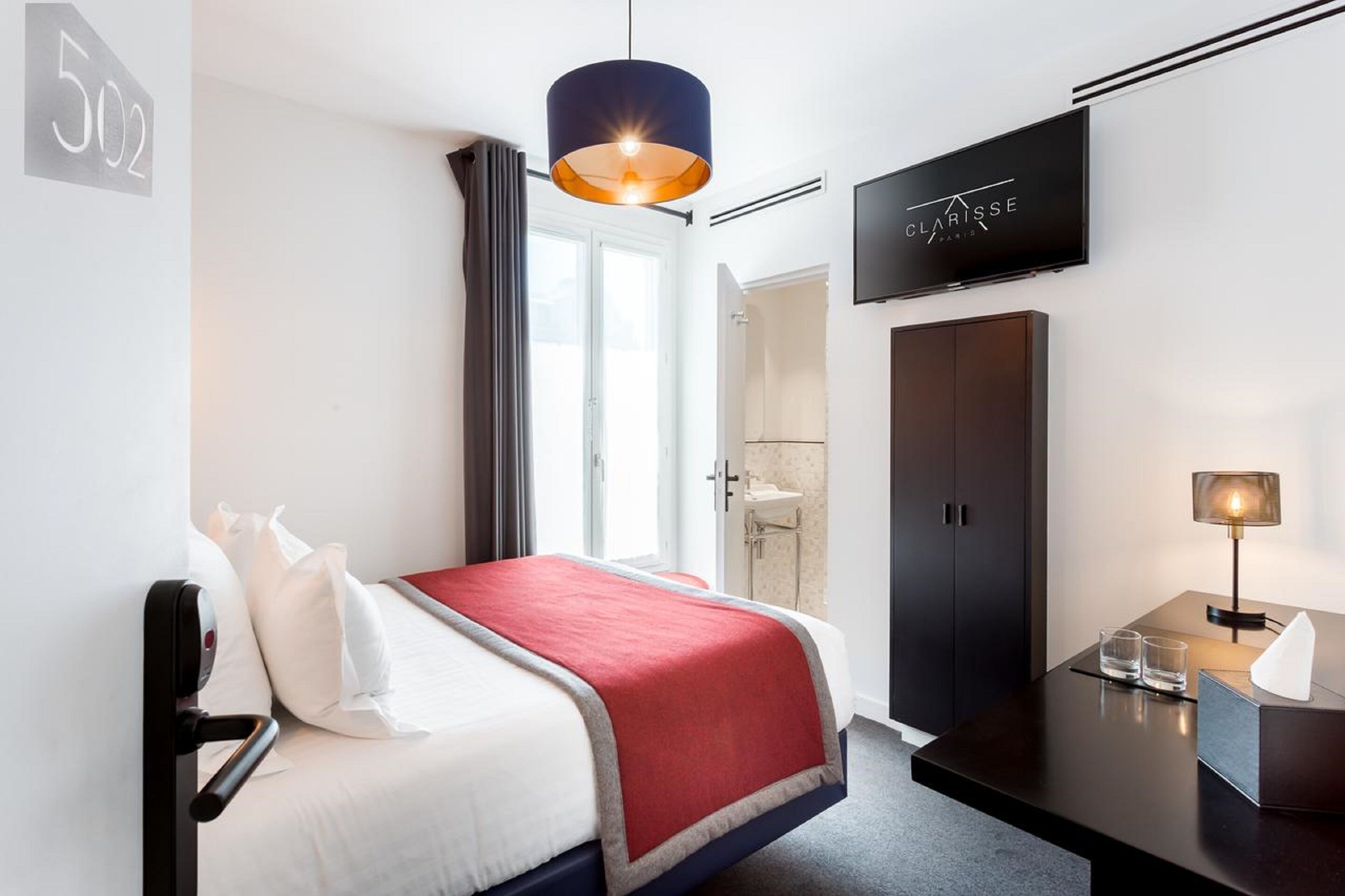 Quarto de hotel moderno com cama, televisão, escrivaninha com luminária e guarda-roupa.