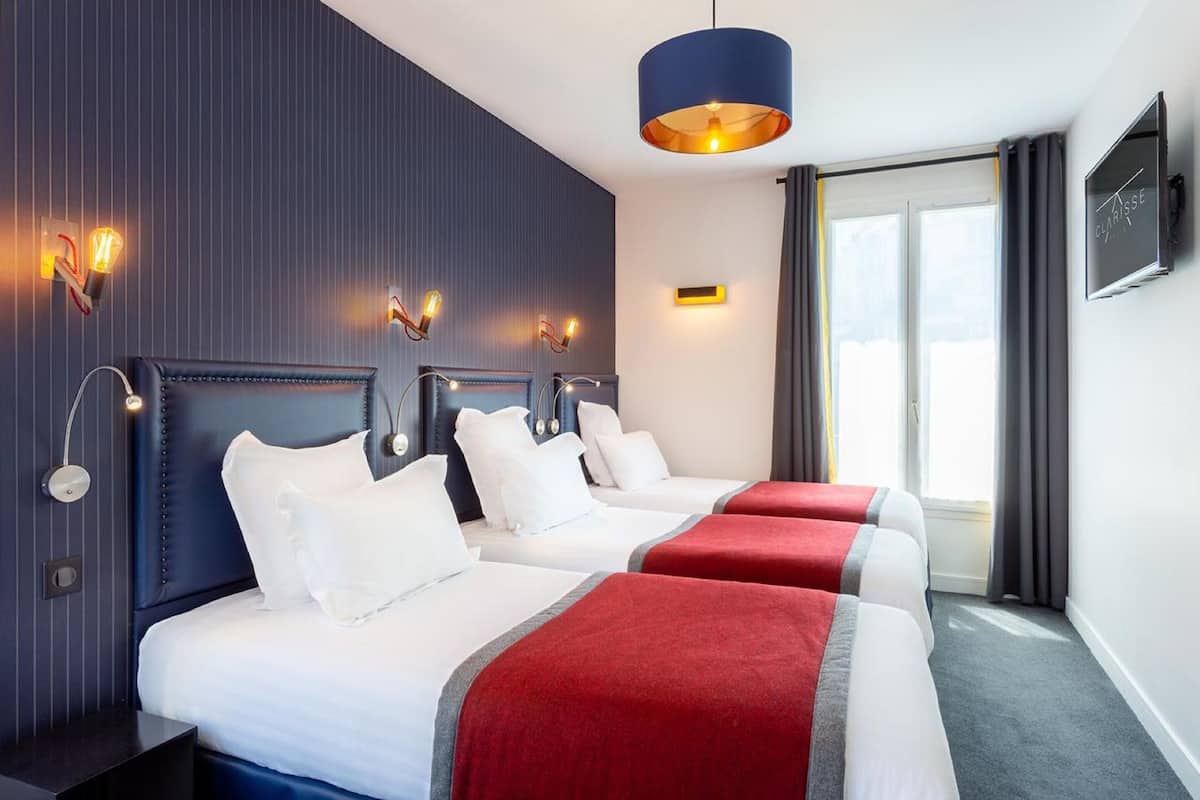 A hotel room with three beds, a large window with curtains, a flat-screen TV, and wall-mounted light fixtures.