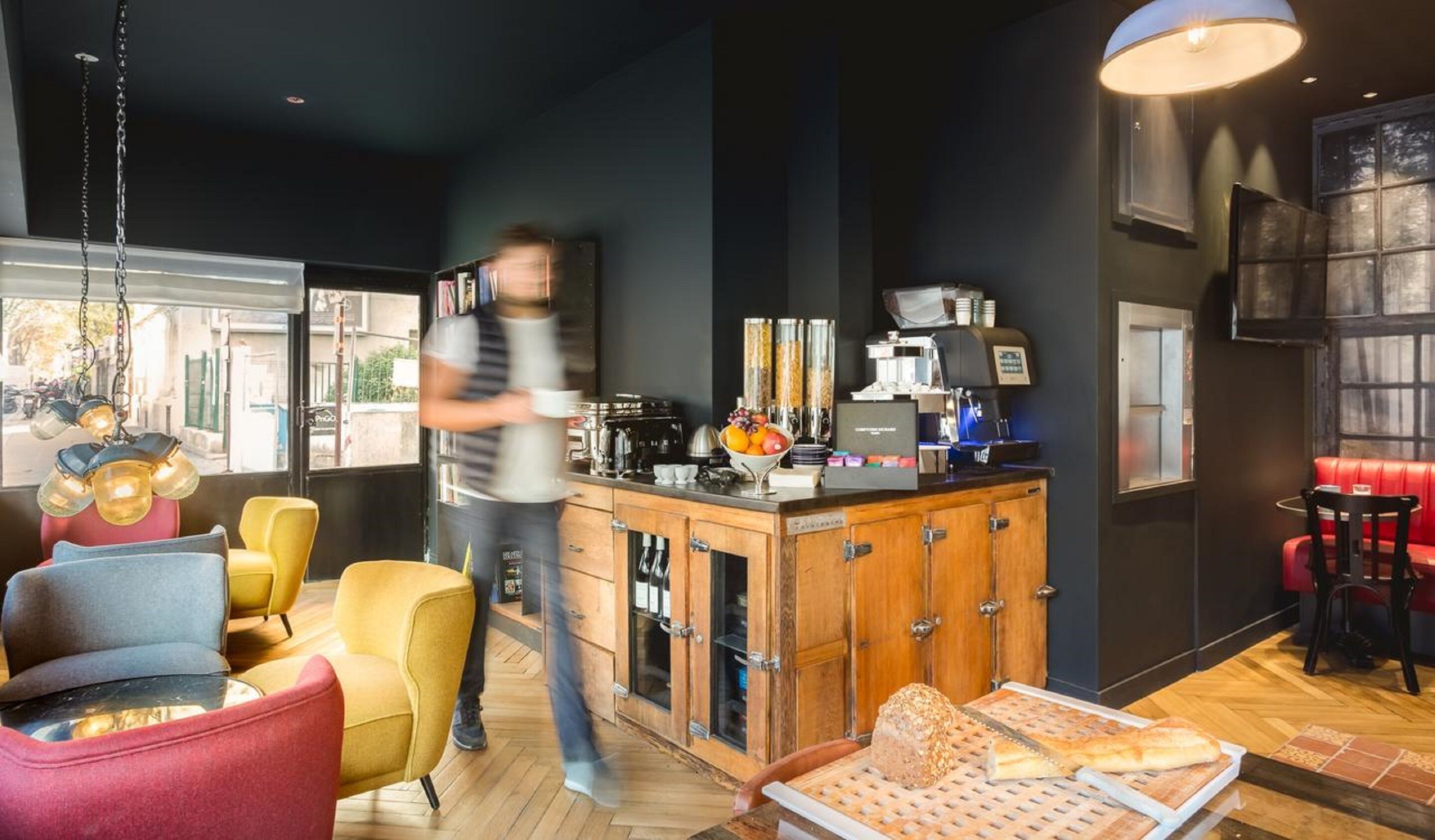 En moderne café med en trædisk, barstole og forskellige siddepladser, herunder en rød sofa og gule stole.