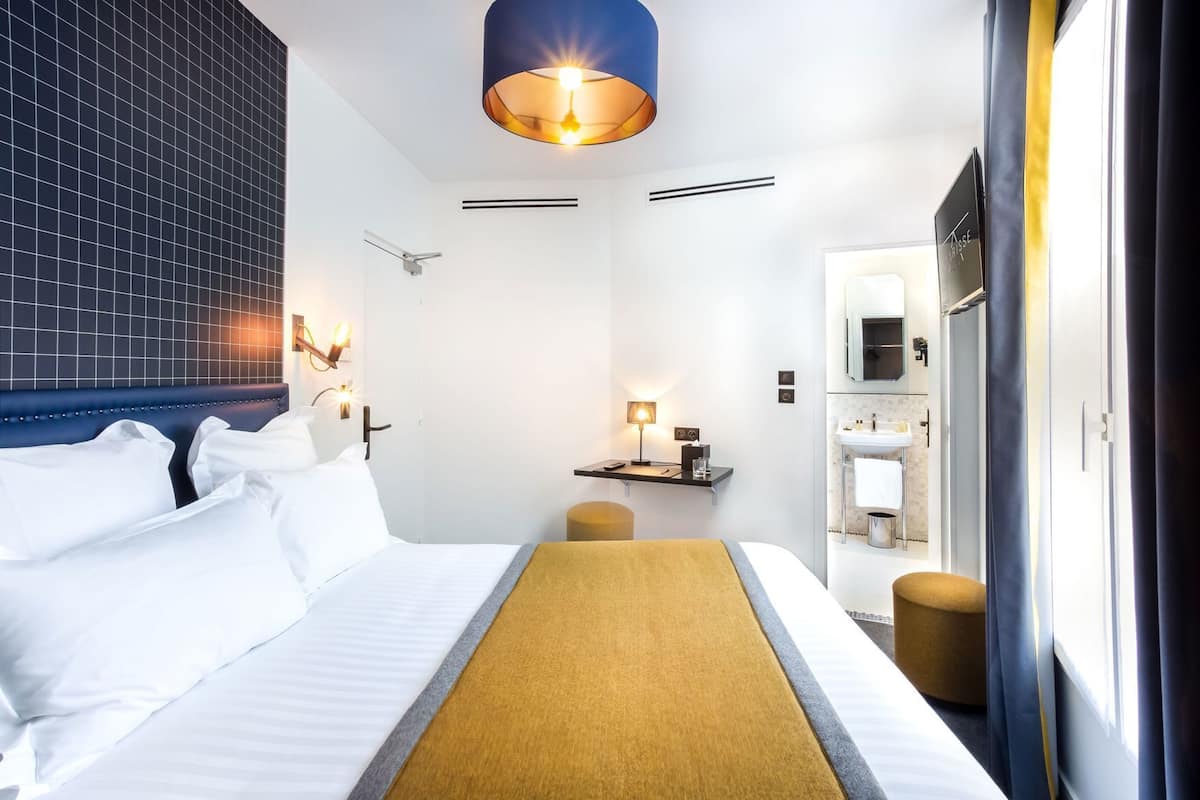A hotel room with a bed, bedside table, lamp, and a view of the outside.