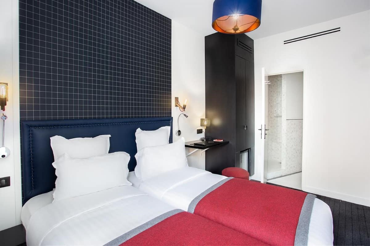 A modern hotel room with a large bed, a dark headboard, a red accent bed runner, a wall-mounted light fixture, and a view of a shower area.