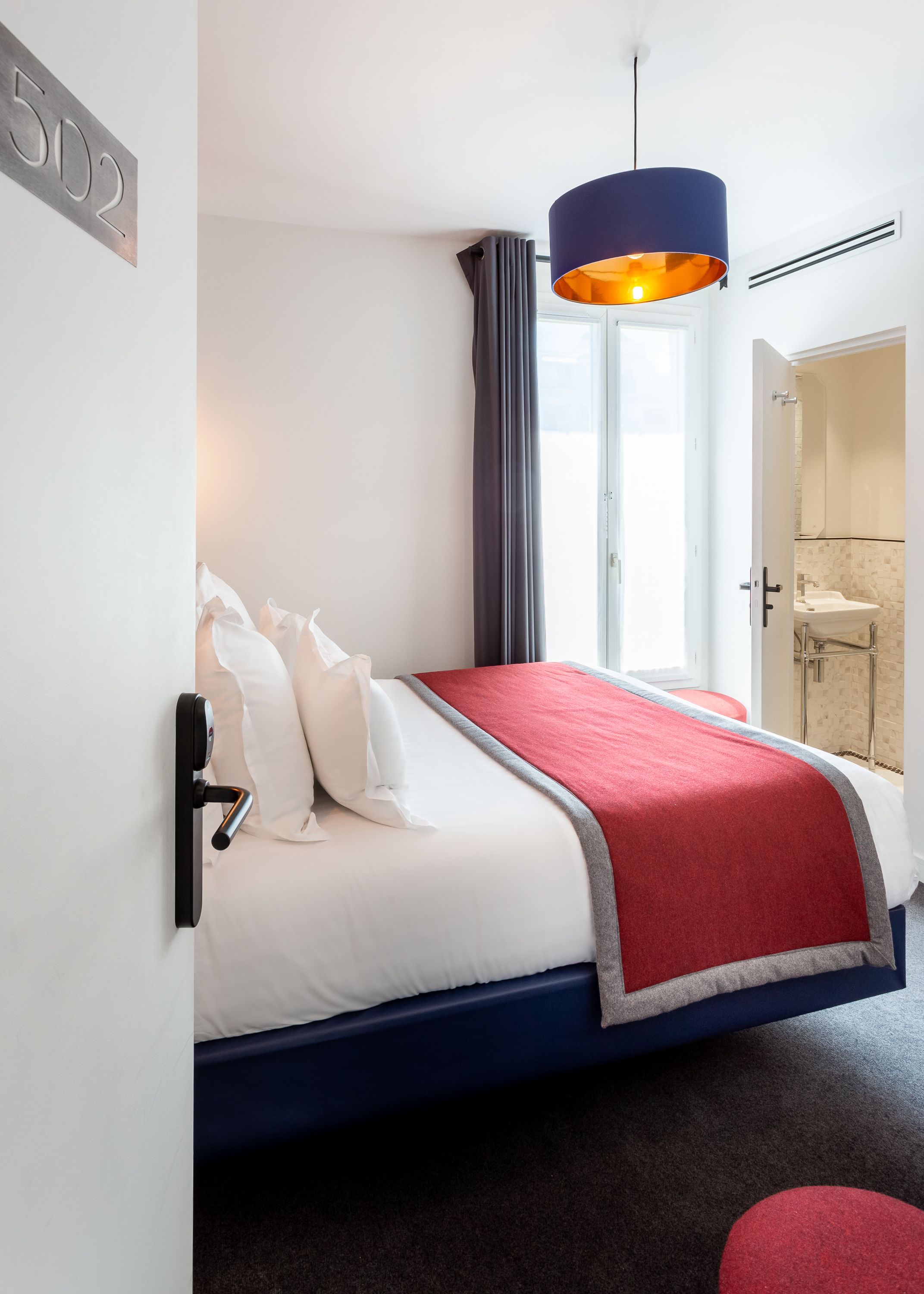 A modern hotel room with a bed, a red and blue bedspread, a large window with curtains, and a bathroom visible through an open door.