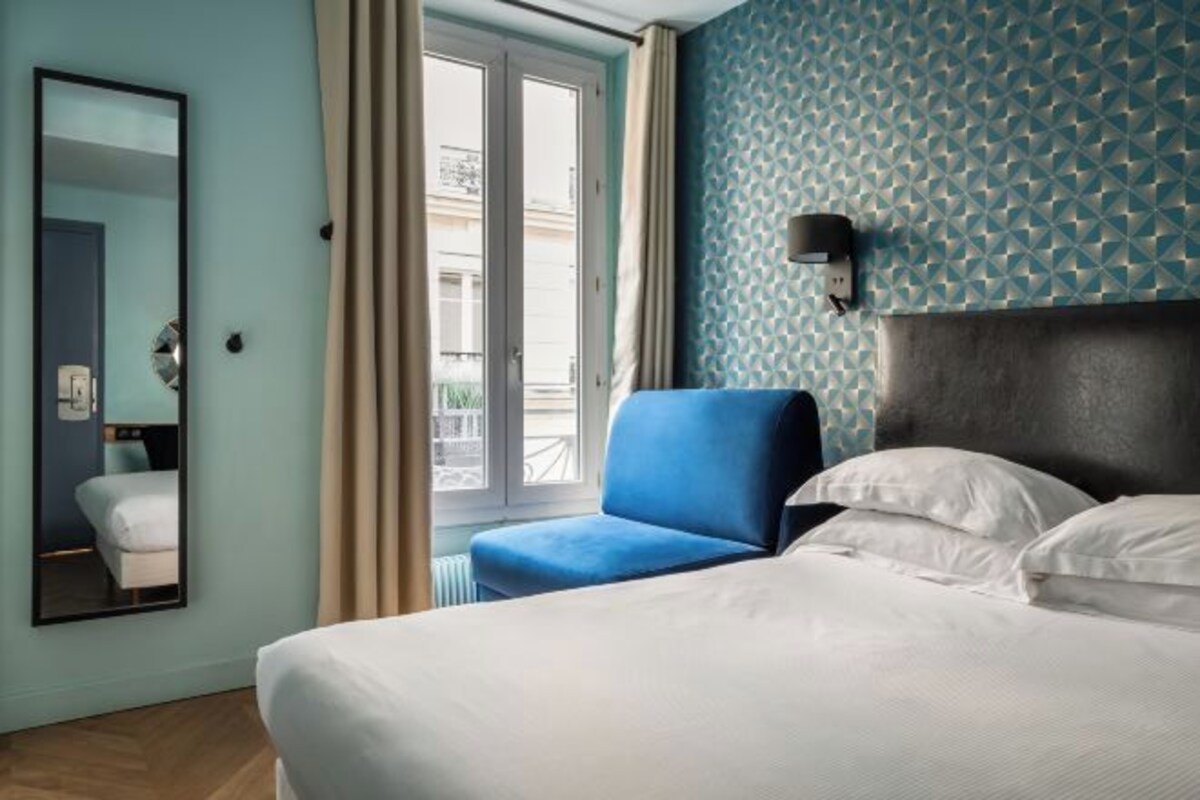 A hotel room with a bed, a blue armchair, a window with curtains, and a wall with a geometric pattern.