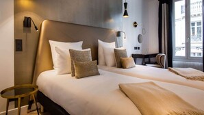 A hotel room with two beds, a desk, and a chair. There are wall-mounted lamps and a window with curtains. - Hotel Royal Opera (Paris)
