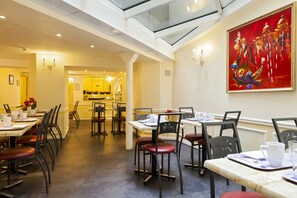 Un restaurant avec de hauts plafonds, une verrière et un grand tableau accroché au mur. Les tables sont dressées avec des nappes blanches et des chaises rouges.