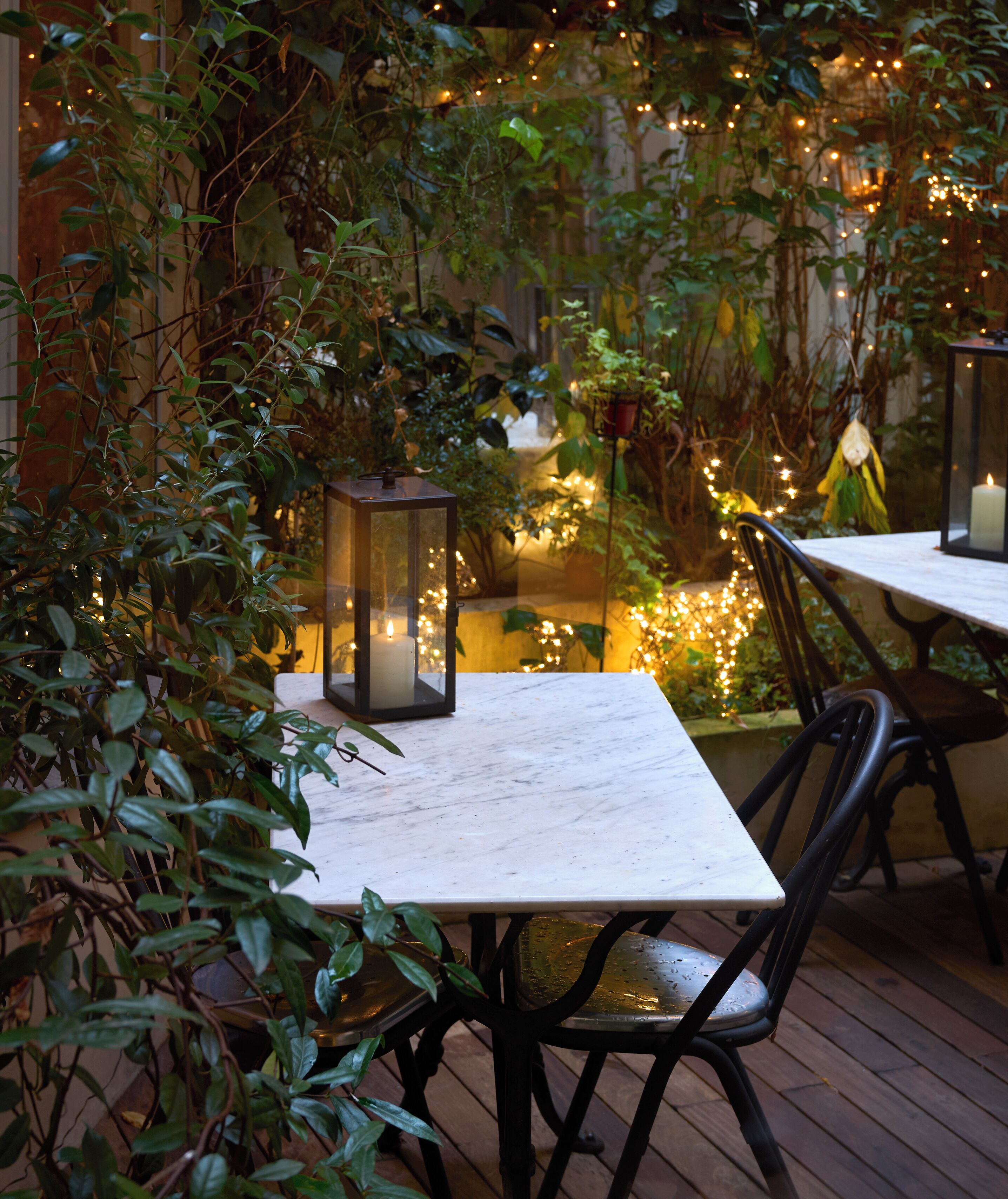 Un espace de restauration en plein air, avec une table et des chaises, entouré de verdure et illuminé de guirlandes lumineuses.