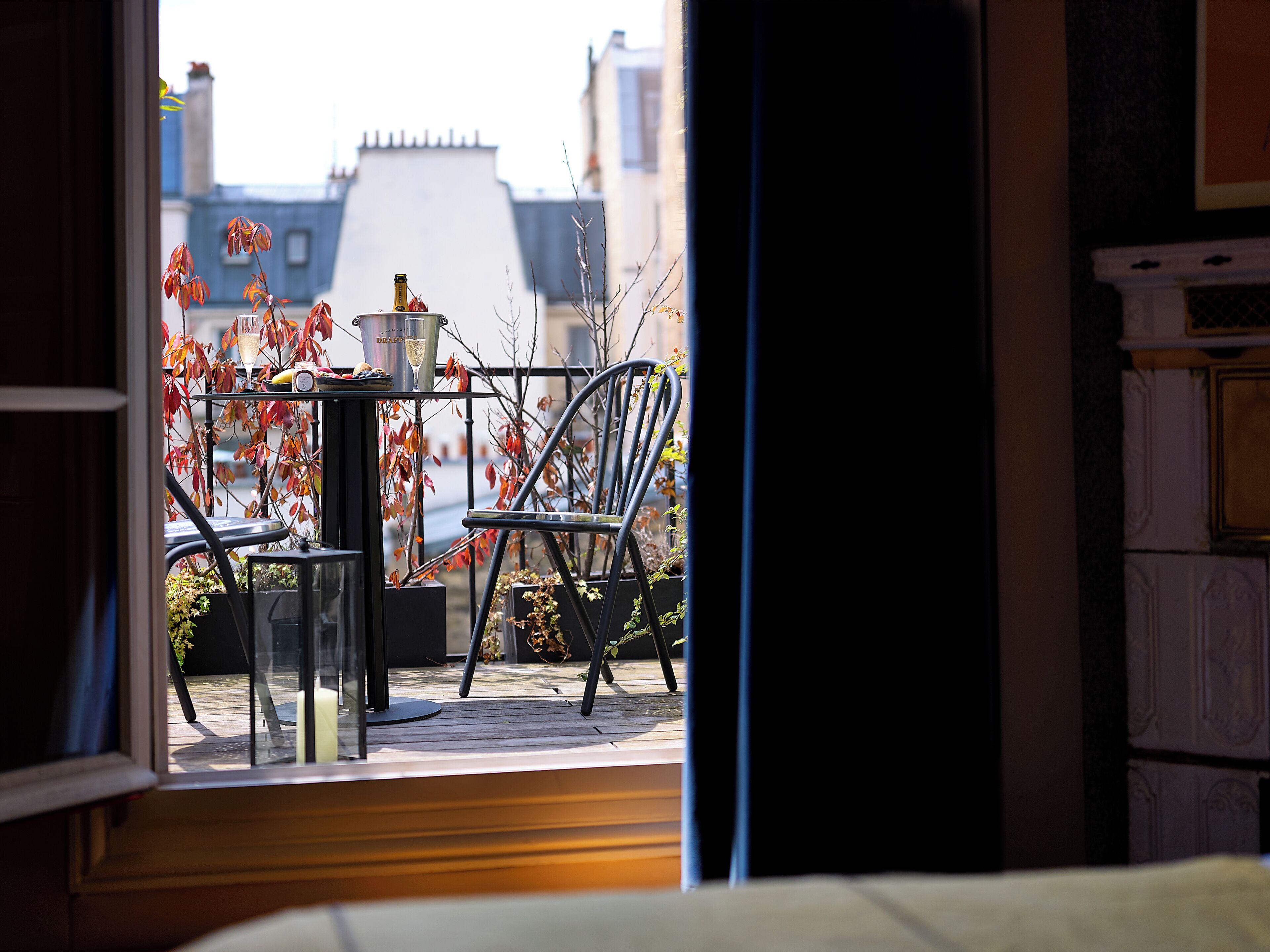 Blick aus einem Zimmer mit Fenster auf einen Balkon, der einen Tisch, Stühle und eine Champagnerflasche zeigt.