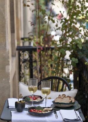 A table set with a plate of vegetables, a bowl of olives, two glasses of white wine, and slices of bread on a balcony with a view of greenery.