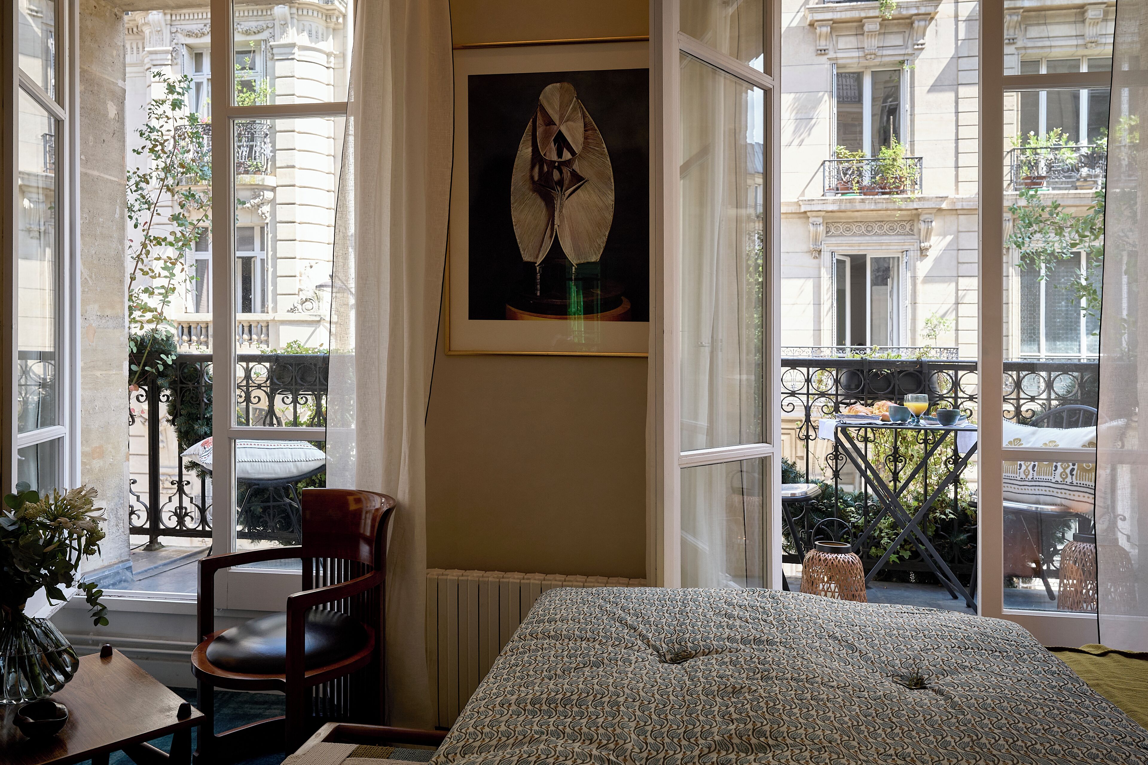 Een kamer met een groot raam, een ingelijste afbeelding, een houten stoel en een bed met een decoratieve spreideken.