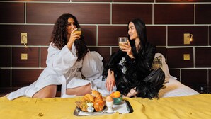 Dos personas sentadas en una cama con una manta amarilla, un plato de comida y un tazón de fruta.