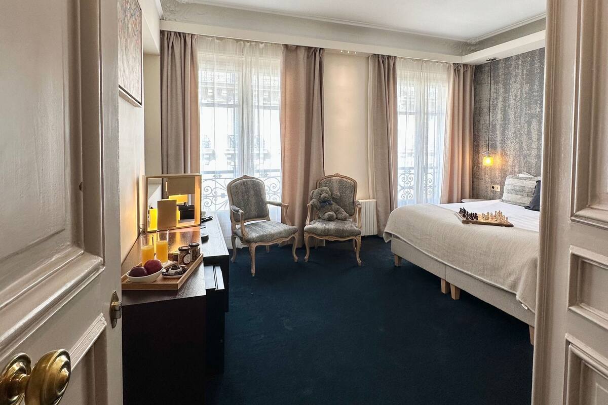 Una habitación de hotel con cama, dos sillones, un escritorio con espejo y ventana con cortinas.