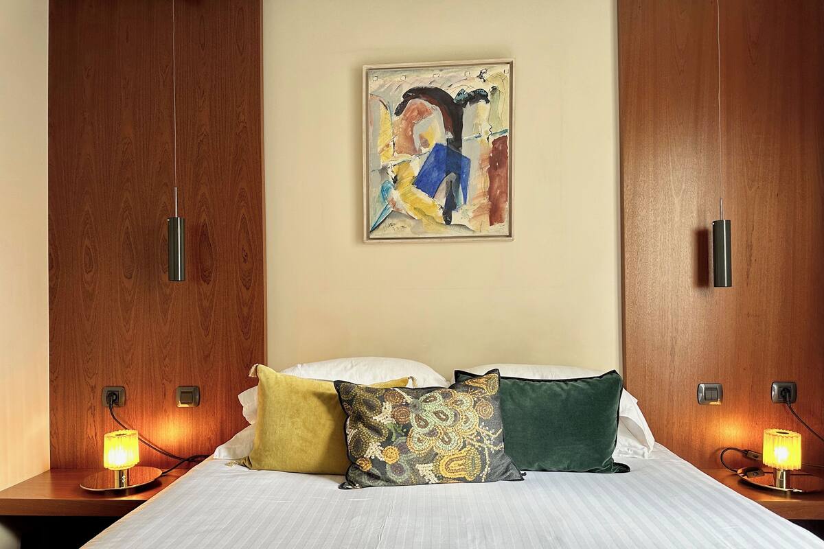 Habitación de hotel con una cama, dos lámparas de noche, un cuadro en la pared y un armario de madera.