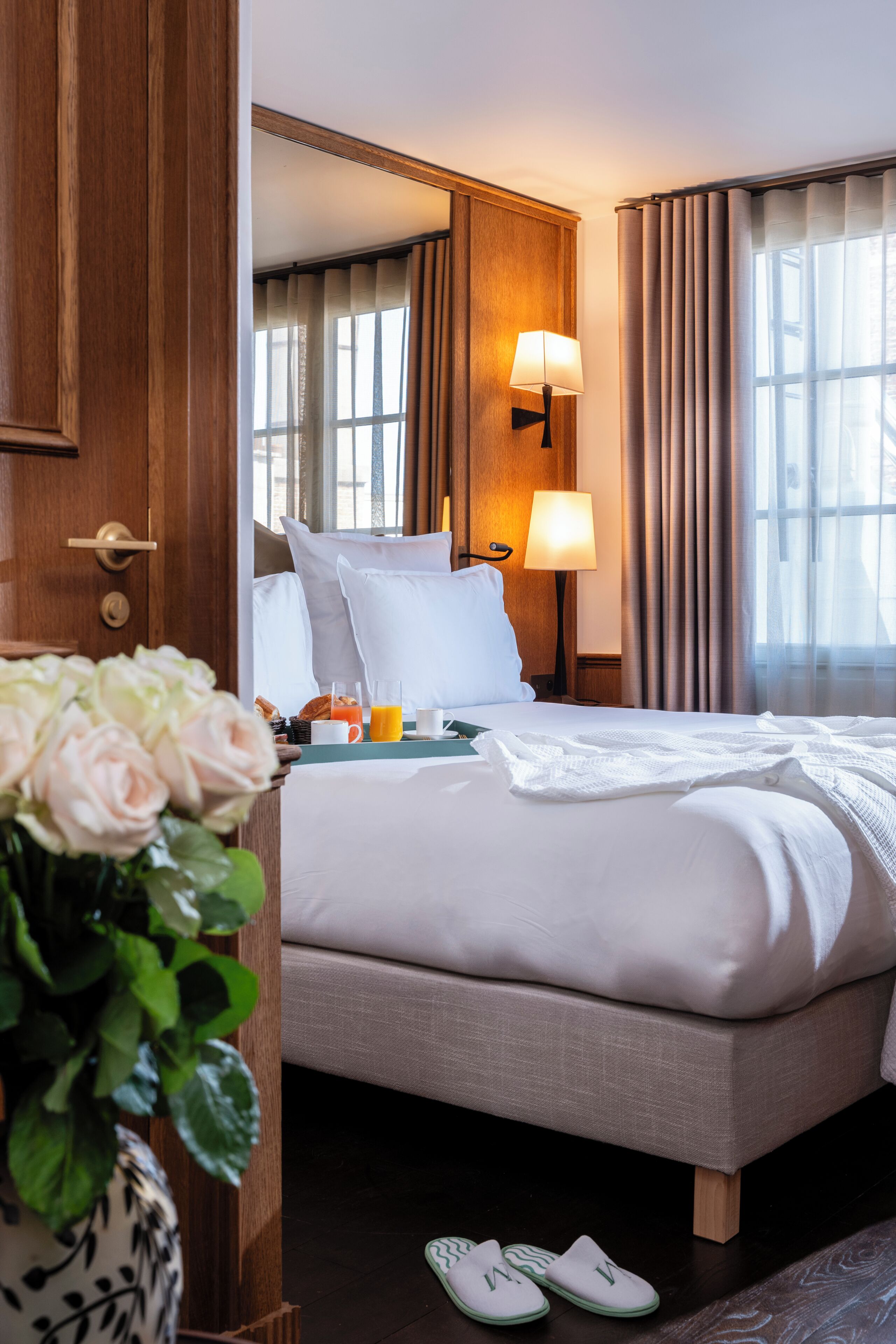 A hotel room with a bed, bedside lamps, a vase of flowers, and a window with curtains.