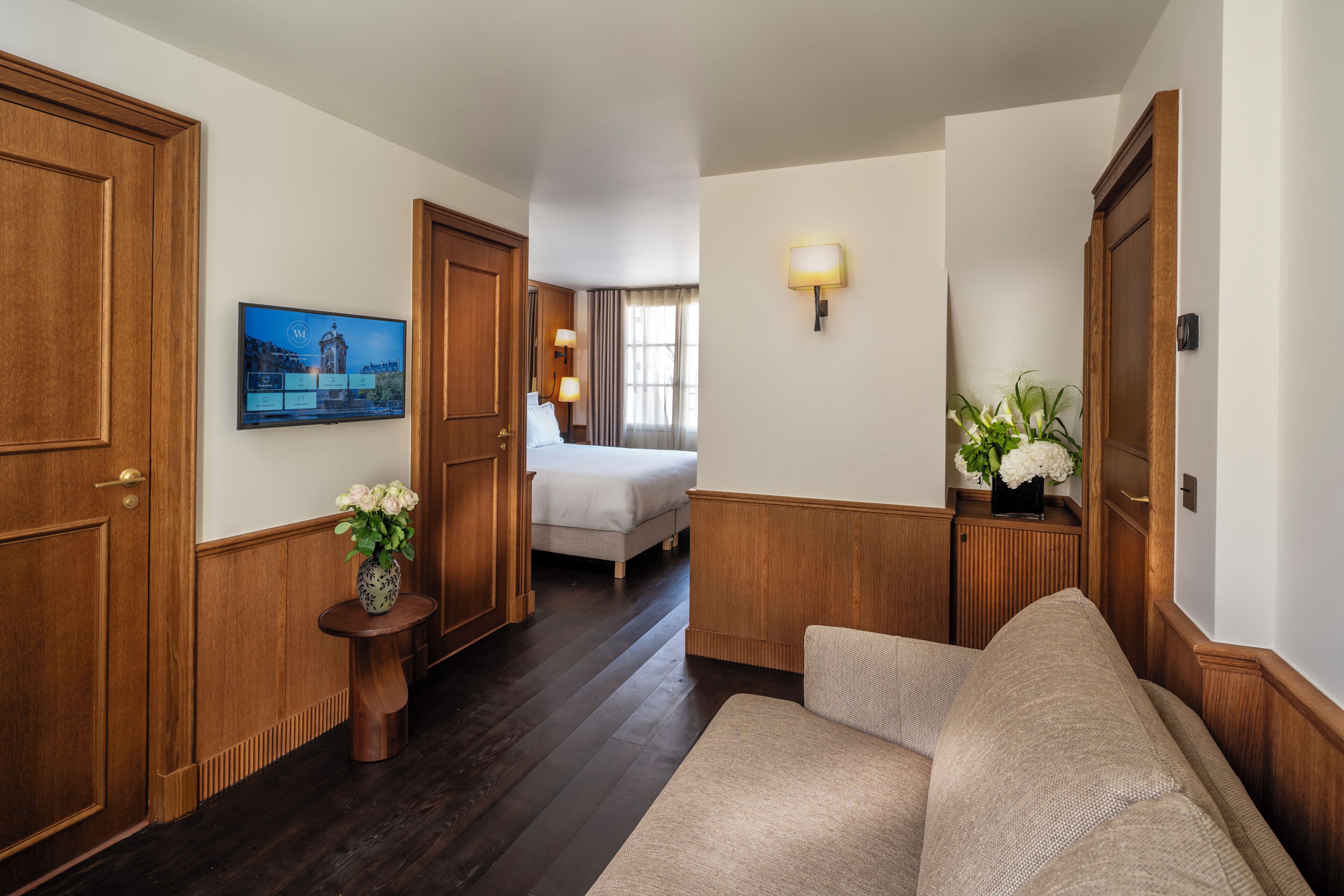 A hotel room with a wooden floor, a sofa, a bed, a small table with a vase, and a wall-mounted TV.