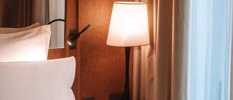 A hotel room with a bed, two wall-mounted lamps, a nightstand with a lamp, and a window with curtains.