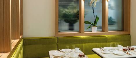 A dining area with green booths and tables, wooden beams on the ceiling, and a window with an orchid plant.