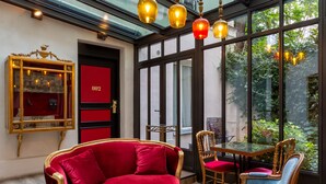 A room with a red velvet sofa, wooden chairs, and a glass table. There are hanging lights and a large window with a view of greenery. - Hôtel Sacha (Paris)