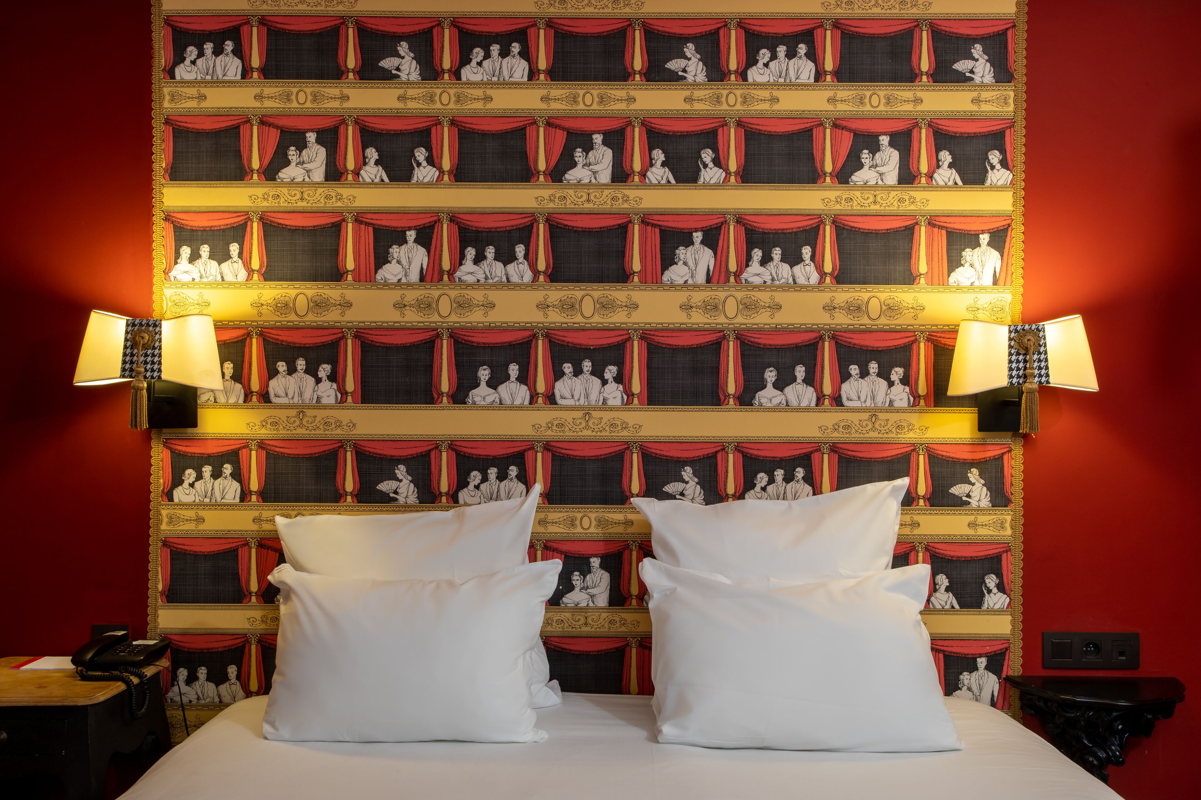 Habitación de hotel con una cama grande, dos almohadas blancas, una mesita de noche con una lámpara y un papel tapiz rojo y dorado con motivos teatrales.