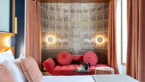 A hotel room with a large bed, a red sofa, a small ottoman, and a coffee table. There are two wall sconces and a large tapestry with intricate designs. - Hôtel Sacha (Paris)