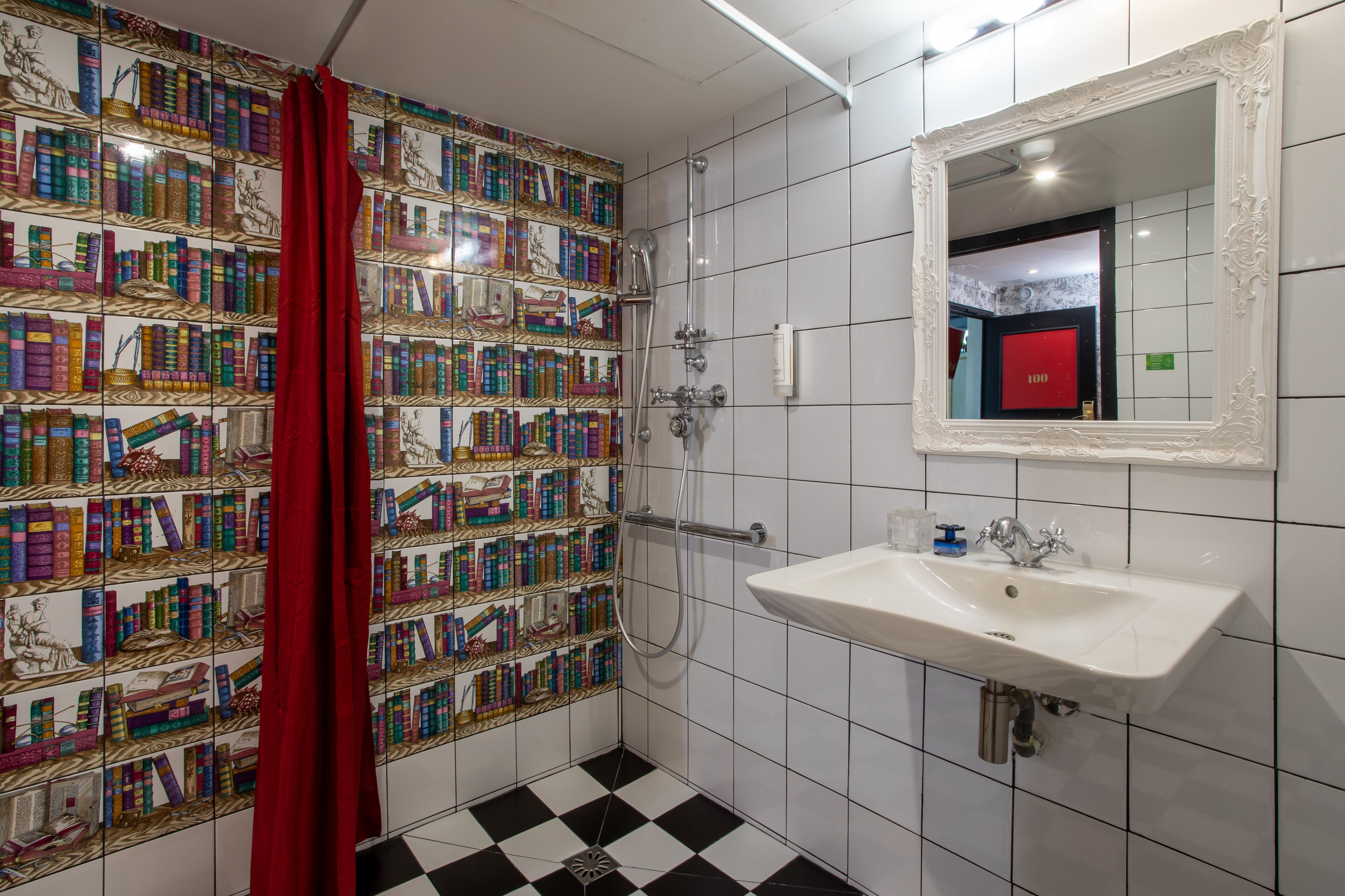 Une salle de bain avec un mur recouvert de carreaux présentant un motif coloré inspiré de livres, un rideau de douche rouge, un lavabo blanc et un miroir avec un cadre orné.