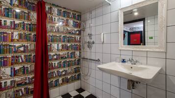Une salle de bain avec un mur recouvert de carreaux présentant un motif coloré inspiré de livres, un rideau de douche rouge, un lavabo blanc et un miroir avec un cadre orné.