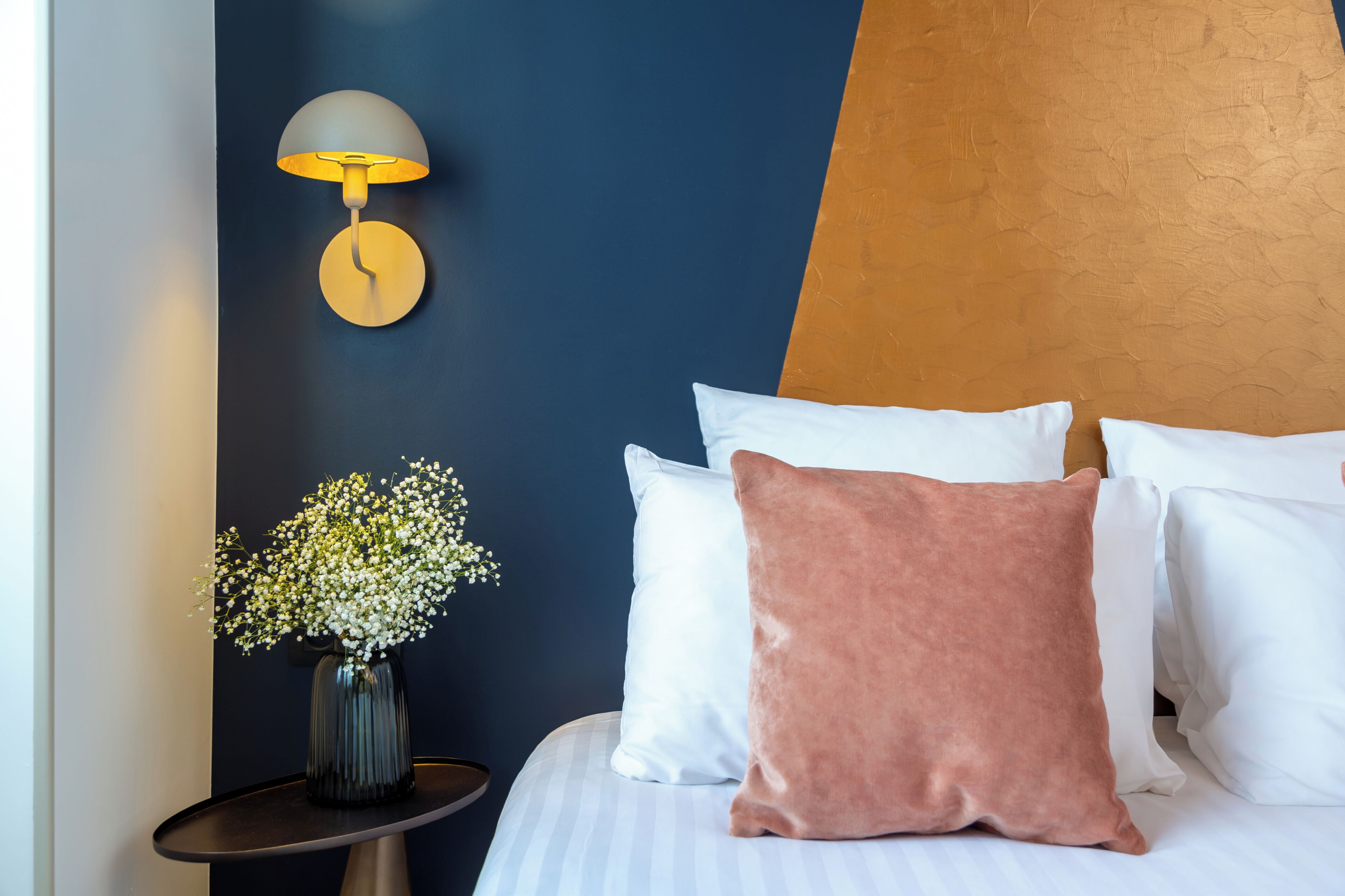 Une chambre à coucher avec un lit, des oreillers, un vase de fleurs, une lampe fixée au mur et une table de chevet.