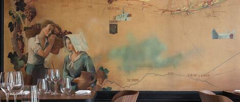 L’intérieur d’un restaurant, avec une grande carte géographique peinte sur le mur, des chaises en bois et des verres à vin sur la table.