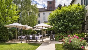 Un patio al aire libre con mesas y sombrillas, rodeado de follaje y un edificio al fondo.
