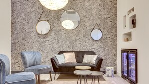 A modern living room with a patterned wallpaper, a dark sofa, two armchairs, a round coffee table, and a small refrigerator. - Hotel Courseine (Courbevoie)