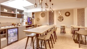 A modern café with a long wooden table, multiple chairs, hanging pendant lights, a refrigerator with beverages, and a coffee machine. - Hotel Courseine (Courbevoie)