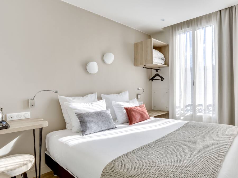 Ein modernes Hotelzimmer mit einem großen Bett, einem Schreibtisch mit Kaffeemaschine, einem Kleiderhaken und einem Fenster mit transparenten Vorhängen.